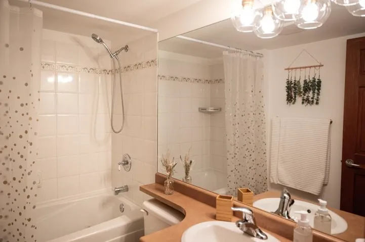 Bathroom featuring a full-size bathtub with shower, white ceramic tile surround with decorative accent border, a vanity with a large mirror, and a built-in light fixture with clear glass globes