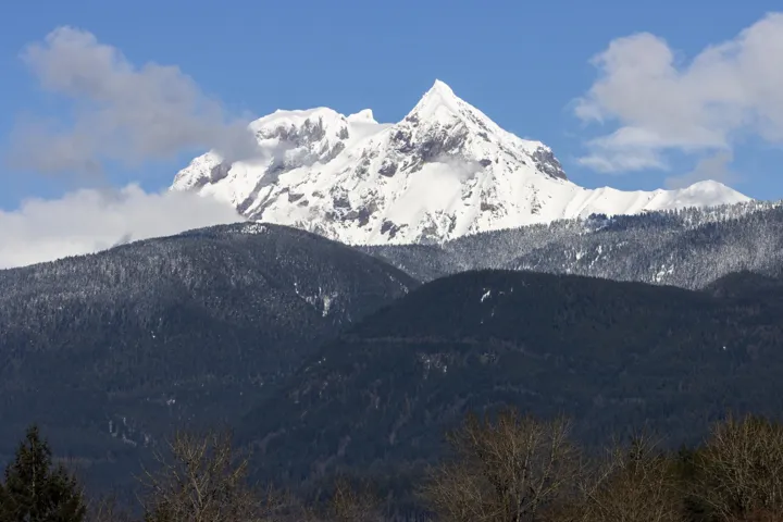 Snow-capped mountain range with a prominent peak, surrounded by lower forested mountains under a blue sky with scattered clouds