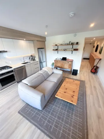 Open-concept living space featuring wood-finish flooring, a contemporary kitchen with stainless steel appliances, white flat-panel cabinetry, and a light-colored countertop with a matching backsplash