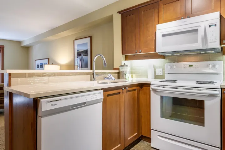 Kitchen with white appliances, brown cabinetry, a peninsula, decorative backsplash, and light stone countertops
