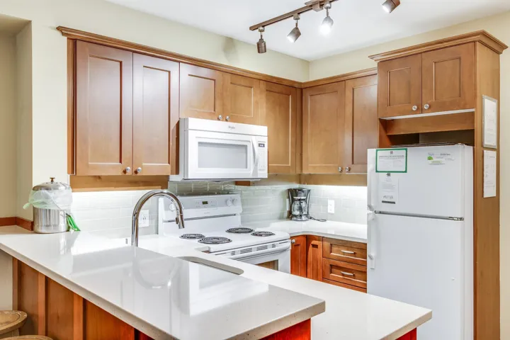 Kitchen featuring white appliances, backsplash, brown cabinetry, light stone countertops, and a peninsula