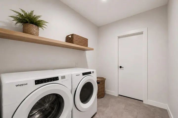 Dedicated laundry area featuring a front-loading washer and dryer, wood-finish floating shelf, and contemporary light gray floor tiles