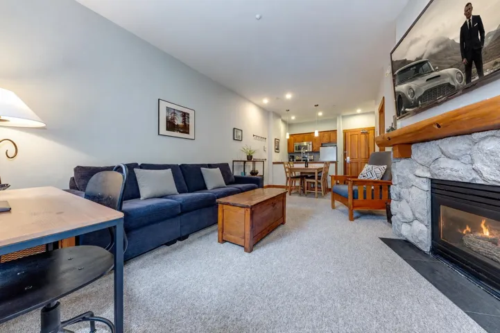 Living area featuring a stone-faced fireplace with a substantial timber mantel, light-toned carpet flooring, recessed lighting, and a partial view of the kitchen and dining area