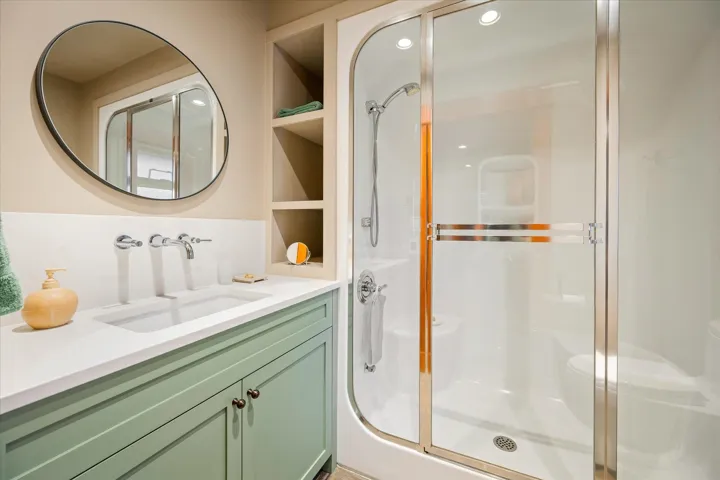Bathroom featuring a walk-in shower with clear glass enclosure, a vanity with a light green cabinet base, a white countertop with an integrated sink, polished chrome wall-mounted faucets, and a round wall mirror