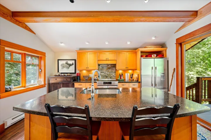 Kitchen featuring a substantial island with a dark countertop and integrated sink, stainless steel appliances, light-toned wood cabinetry, wood-finish flooring, and exposed wood beams