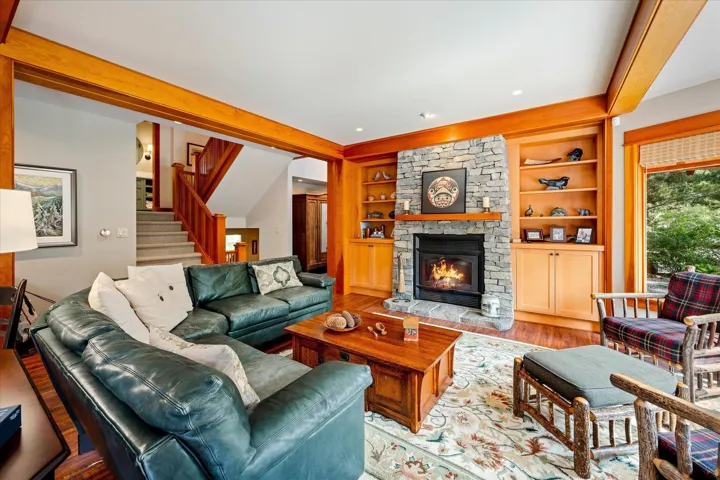 Living area featuring a floor-to-ceiling stone fireplace with a wood mantel, built-in shelving, hardwood flooring, a large window, and exposed wood beams