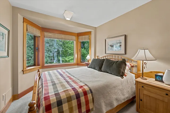 Bay window with natural wood trim framing exterior foliage, beige wall paint, light-colored carpet flooring, and a flush mount ceiling light