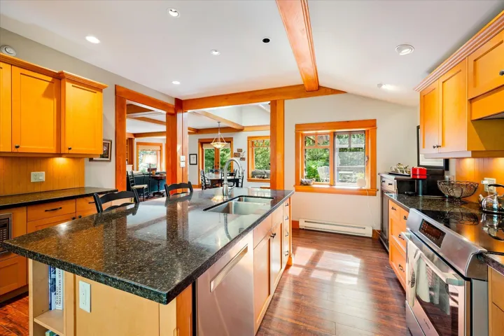 Kitchen featuring wood-finish flooring, wood cabinetry with dark hardware, a center island with an undermount sink, dark speckled countertops, and stainless steel appliances