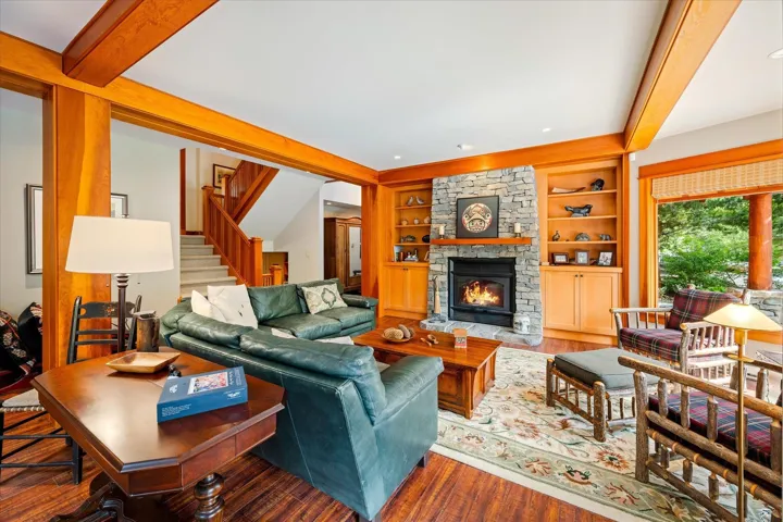 Spacious living area featuring exposed wood beams, a stone-clad fireplace with built-in shelving, dark wood-finish flooring, and a broad window with a natural wood frame
