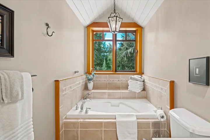 Soaking tub with tiled surround and integrated steps, wood-framed window, vaulted ceiling with shiplap finish, and decorative pendant lighting