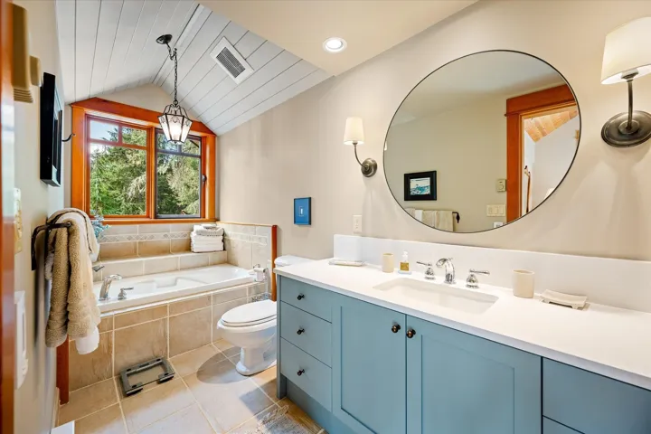 Bathroom featuring a built-in tub with tile surround, a large window with wood trim, sloped plank ceiling, and a vanity with a white countertop and blue cabinetry