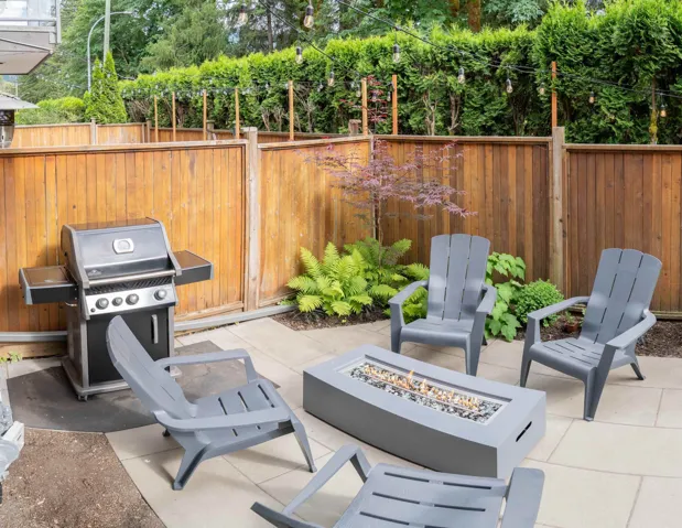 Patio area featuring light-toned paving stones, a built-in fire pit, and a tall wood fence