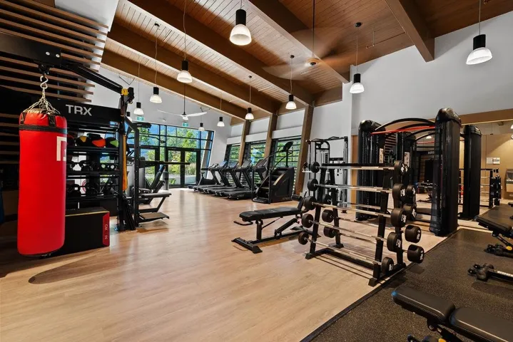 Spacious fitness center featuring vaulted ceilings with exposed wood beams, light wood-finish flooring, floor-to-ceiling windows, and extensive exercise equipment