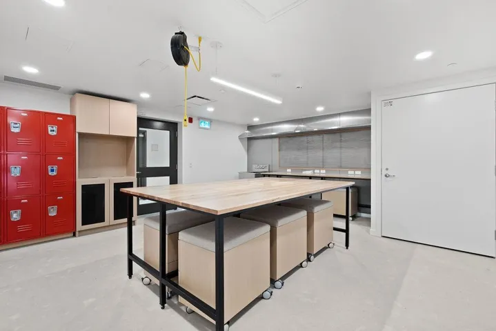 Spacious interior featuring a large central wood-finish table with integrated seating, extensive red locker storage, light-toned built-in cabinetry, recessed lighting, and a durable light-colored floor