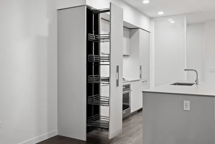 Modern kitchen featuring a pull-out pantry with wire shelving, recessed lighting, wood-finish flooring, a stainless steel wall oven, and a kitchen island with an integrated sink and gooseneck faucet