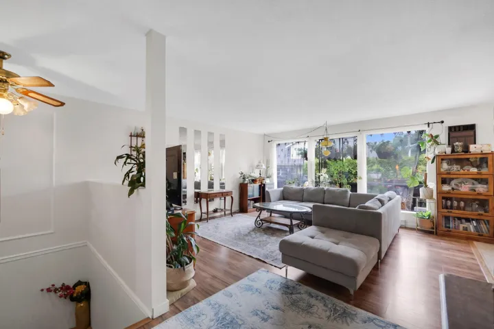 Bright living area featuring large windows, wood-finish flooring, white walls, a ceiling fan with light, and a column detail