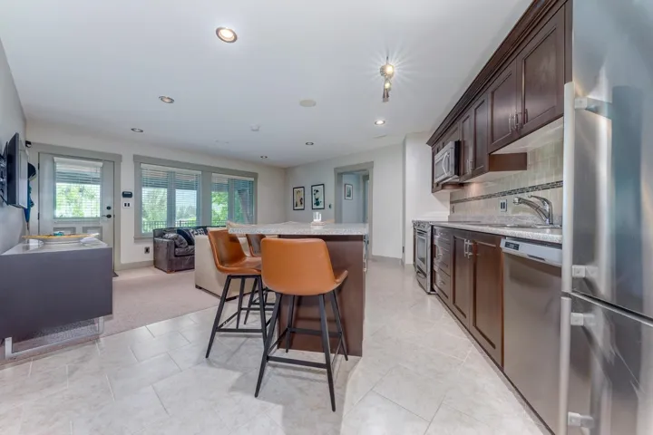 Integrated kitchen and living area featuring stainless steel appliances, dark wood-finish cabinetry, a tile backsplash, recessed lighting, and large windows