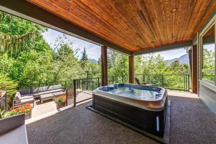 Covered outdoor patio featuring a built-in hot tub, wood-finish ceiling, exposed aggregate flooring, and black metal railings with glass panels