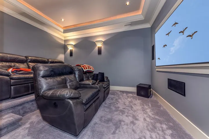 Dedicated media room featuring a large projection screen, built-in sound system, tiered seating platform, recessed ceiling lights, and coved ceiling lighting