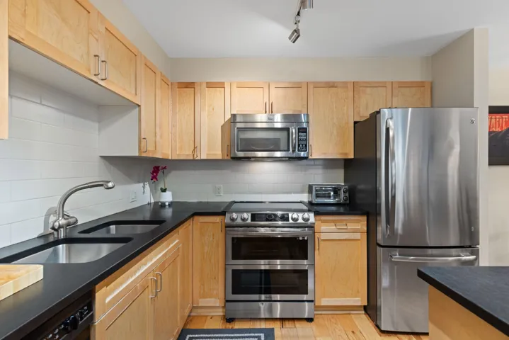 Kitchen with appliances with stainless steel finishes, tasteful backsplash, light brown cabinets, light wood-style floors, and dark stone countertops