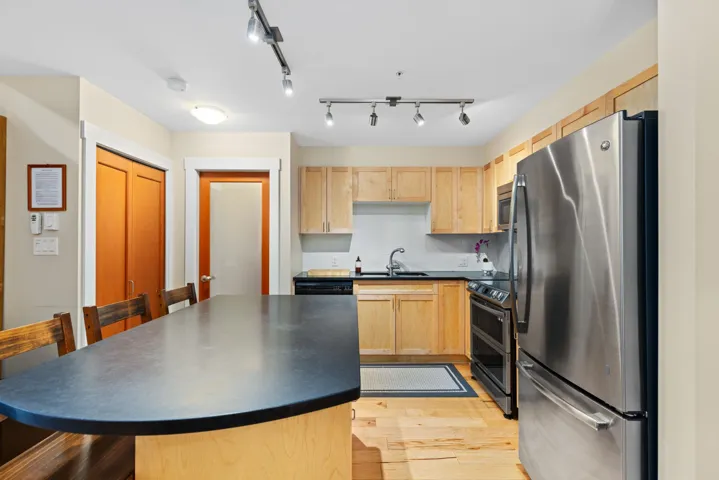 Kitchen with stainless steel appliances, backsplash, light brown cabinetry, light wood-style flooring, and dark countertops