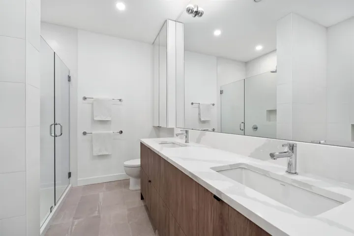 Dual undermount sinks with chrome faucets, wood-finish vanity with ample storage, large integrated mirror, and glass-enclosed shower with white tile surround