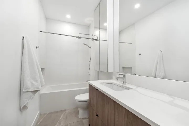 Bathroom featuring a white bathtub with a shower, a wood-finish vanity with an integrated sink, a large mirrored medicine cabinet, and light-toned floor tiling