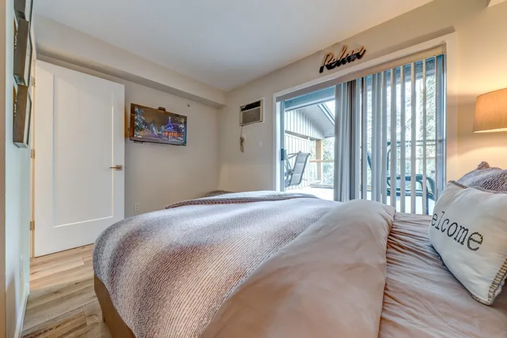 Bedroom featuring a sliding glass door to an exterior balcony, wood-finish flooring, a wall-mounted television, and a window-mounted air conditioning unit