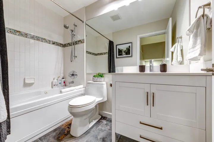 Bathroom featuring a white vanity with shaker-style cabinetry, a built-in bathtub with shower, white subway tile surround with decorative accent, and dark-toned flooring