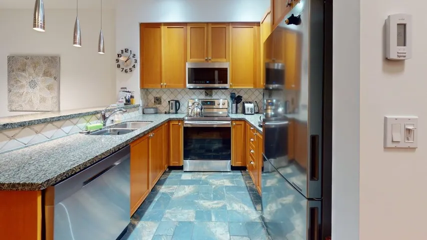 Kitchen with stainless steel appliances, light stone counters, backsplash, decorative light fixtures, and brown cabinetry