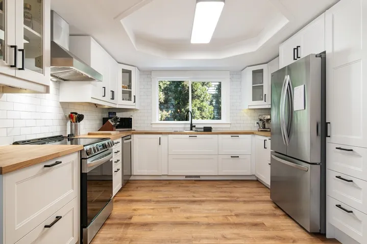 Kitchen featuring butcher block counters, a tray ceiling, stainless steel appliances, light wood-style flooring, and wall chimney exhaust hood