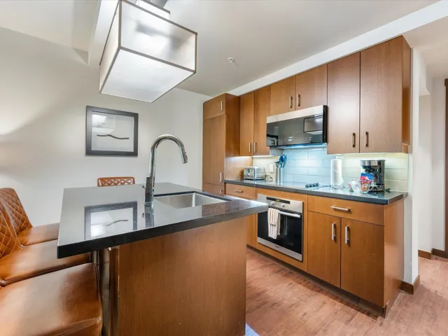 Kitchen featuring a kitchen island with sink, backsplash, stainless steel appliances, brown cabinetry, and dark wood finished floors