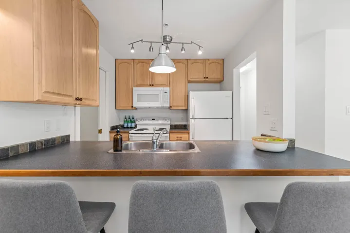 Kitchen with dark countertops, a peninsula, white appliances, and a kitchen bar