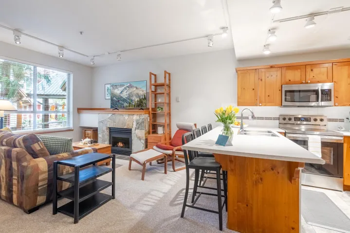 Open-concept living area featuring a stone-clad fireplace, large windows, wood-finish cabinetry, stainless steel appliances, and a kitchen island with seating