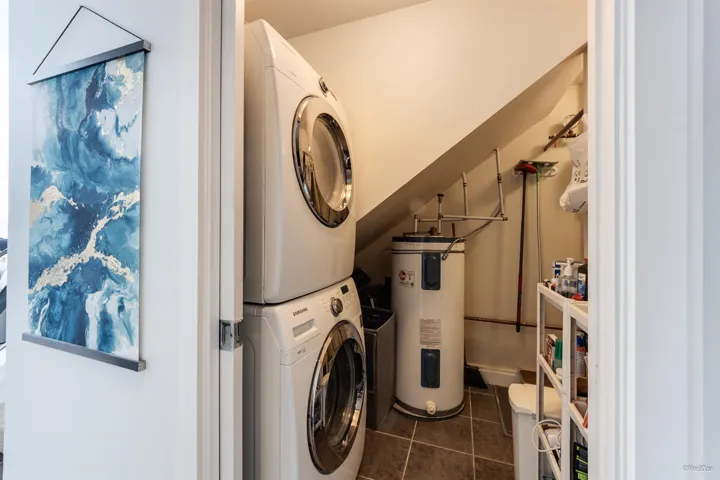 Washroom with electric water heater, stacked washer / drying machine, laundry area, and dark tile patterned flooring