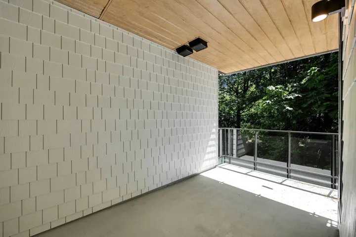 Covered balcony featuring a wood-finish ceiling, textured white wall panels, and a glass-paneled railing with metal posts