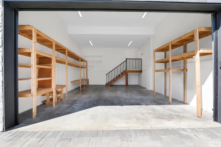 Spacious garage featuring a concrete floor, built-in wooden shelving, a staircase with black railings, and recessed lighting