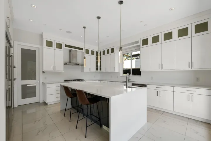Bright kitchen featuring a waterfall island with a dark base, white cabinetry with upper glass-front accents, stainless steel range hood, stone-finish countertops, and large format tile flooring