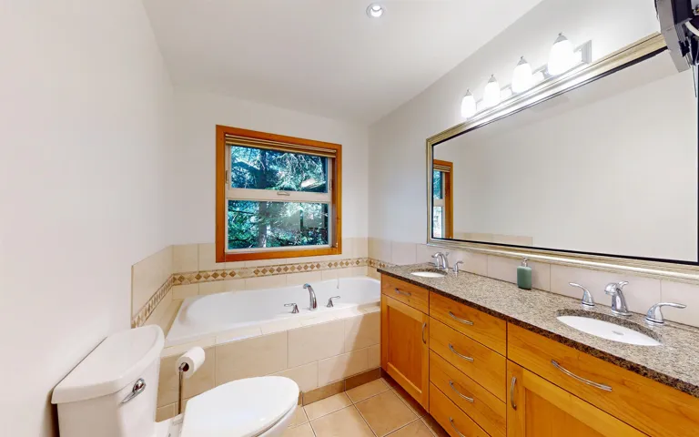 Bathroom featuring a built-in soaking tub with tiled surround, a window with wood trim, a double vanity with stone countertop and integrated sinks, and a large wall-mounted mirror with overhead lighting