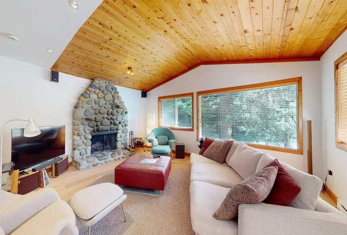 Living area featuring a floor-to-ceiling river stone fireplace, wood-plank vaulted ceiling, and multiple windows with wood trim