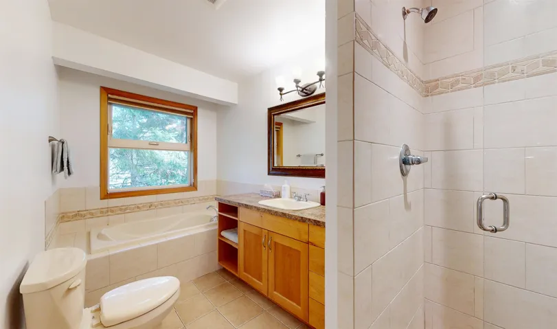 Full bathroom featuring a tiled shower, a soaking tub with tiled surround, a wooden-framed window, a vanity with a light-toned countertop, and a mirrored medicine cabinet with integrated lighting