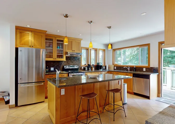 Kitchen featuring light wood cabinetry, stainless steel appliances, a dark tile backsplash, and a central island with a granite-style countertop