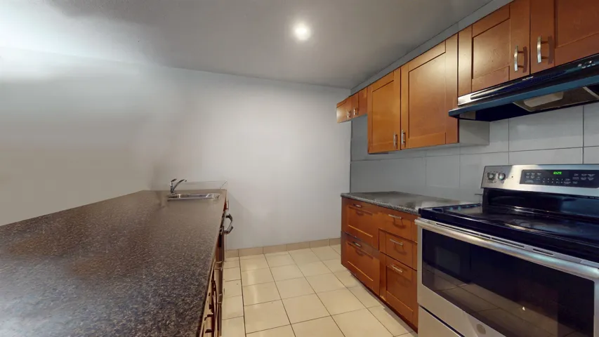 Kitchen featuring stainless steel range with electric stovetop, under cabinet range hood, dark countertops, brown cabinetry, and backsplash