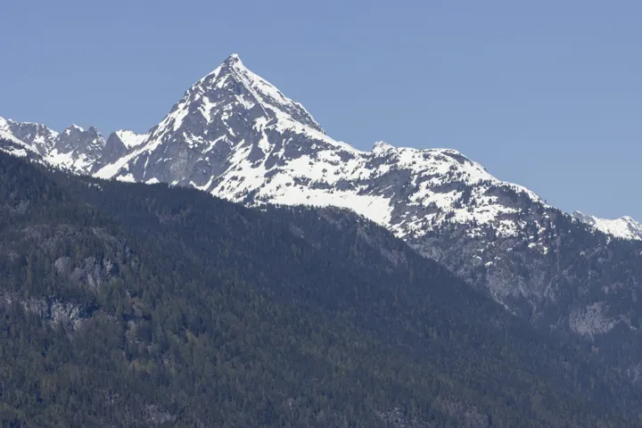Expansive mountain backdrop with snow-capped peaks, dense forest surroundings, and a residential area with varied rooflines