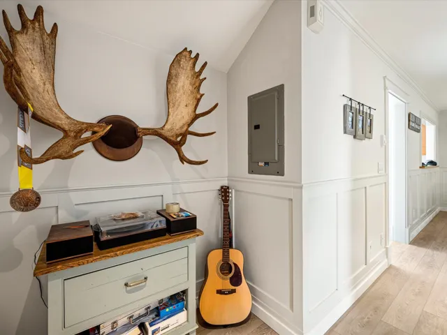 Hallway with electric panel, light wood-type flooring, crown molding, a decorative wall, and a wainscoted wall