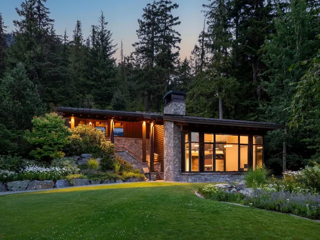 Back of house at dusk featuring a lawn, a chimney, stairs, stone siding, and a view of trees