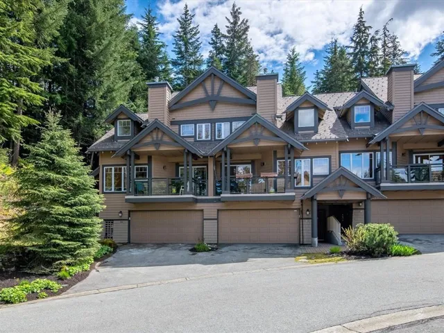Townhouse exterior with multiple dormer windows, covered balconies with glass railings, two integrated garages, and a prominent front entrance with a covered porch