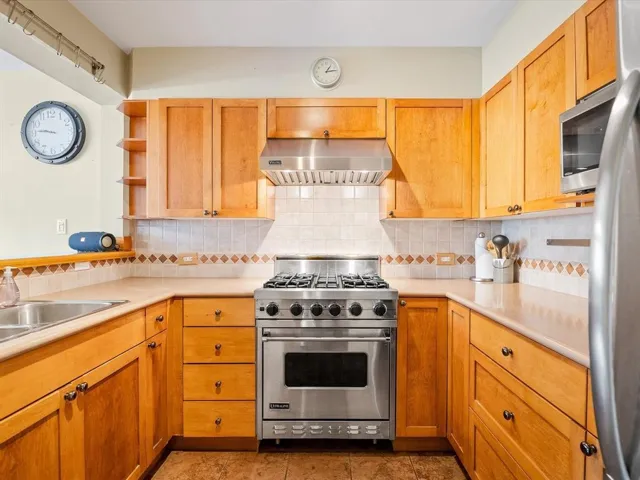 Kitchen featuring a stainless steel gas range with a matching range hood, light wood cabinetry, a tile backsplash with a decorative border, and light-toned countertops