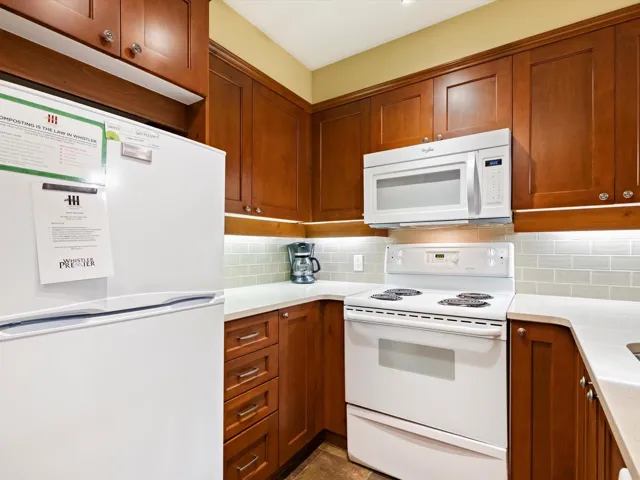 Kitchen with white appliances, brown cabinetry, backsplash, and light stone counters