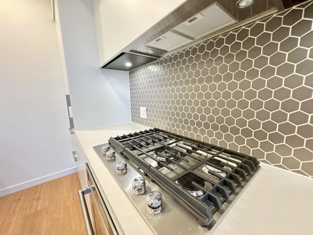 Kitchen view of wall chimney exhaust hood, stainless steel gas stovetop, backsplash, light stone countertops, and light wood-style floors